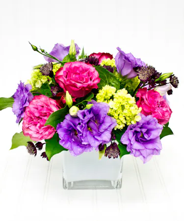 A glass cube vase, filled with lavender roses, and hydrangea accented with bright purple status.