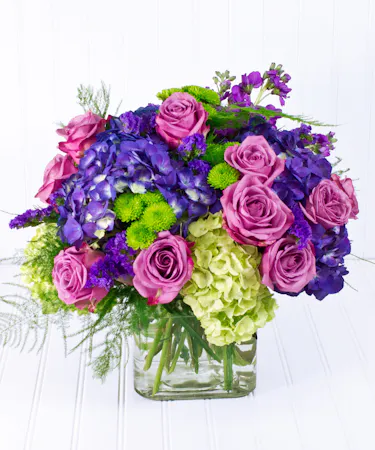 A glass cube vase, filled with lavender roses, and hydrangea accented with bright purple status.